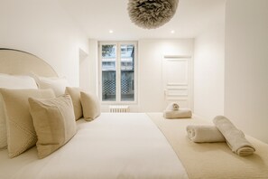 Et minimalistisk soveværelse med en stor seng, en radiator og et vindue med udsigt til bygninger.