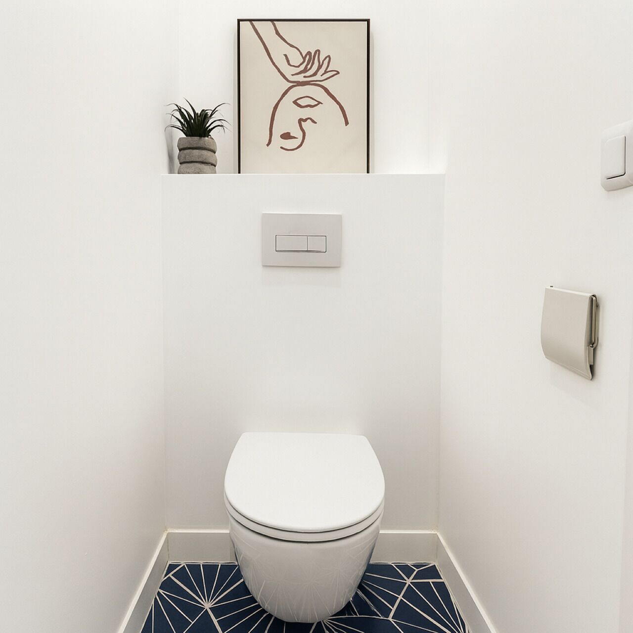 A compact bathroom with a white toilet, a wall-mounted light switch, and a framed artwork above the toilet.