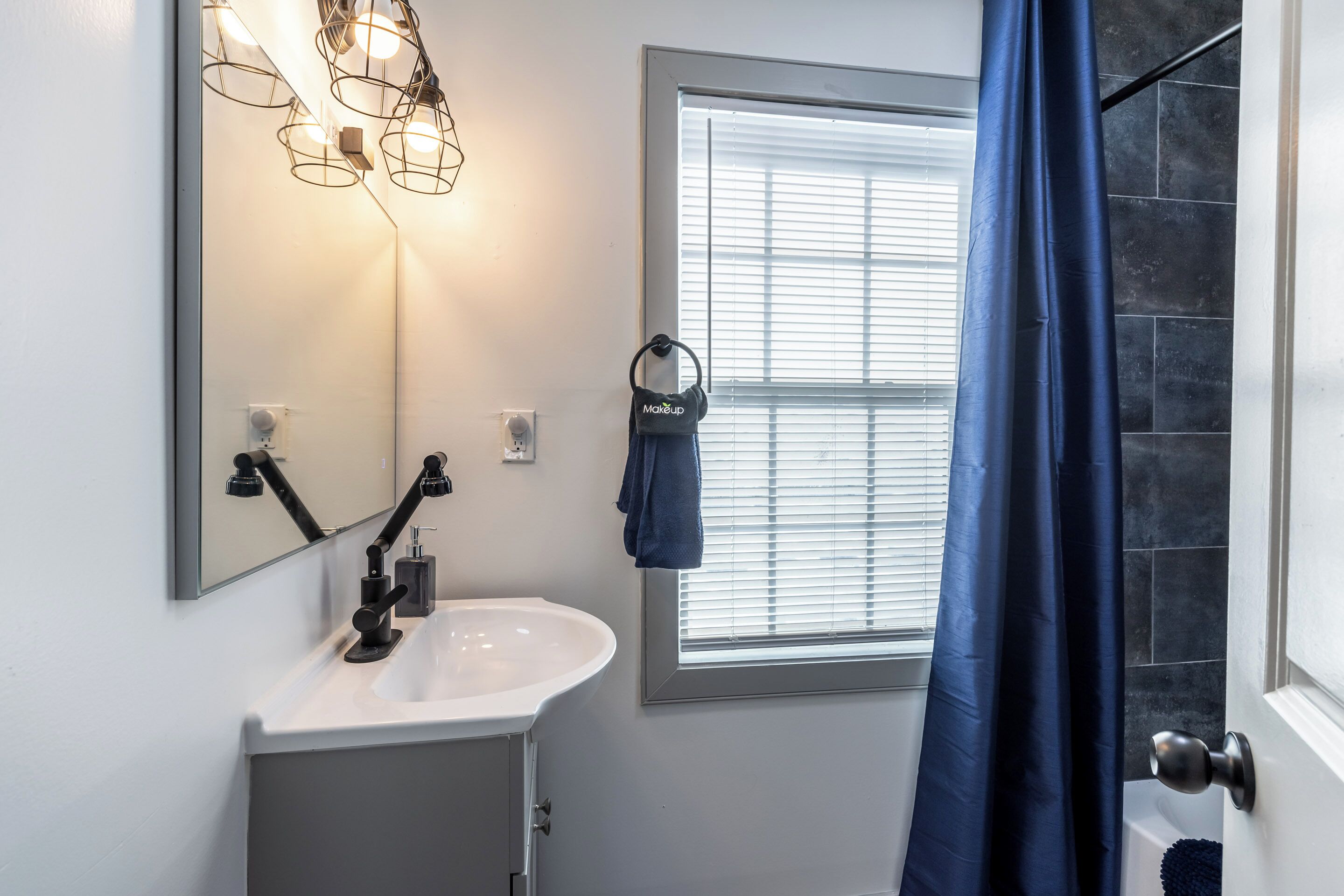 Un baño con lavamanos blanco, un espejo y una ventana con persianas.
