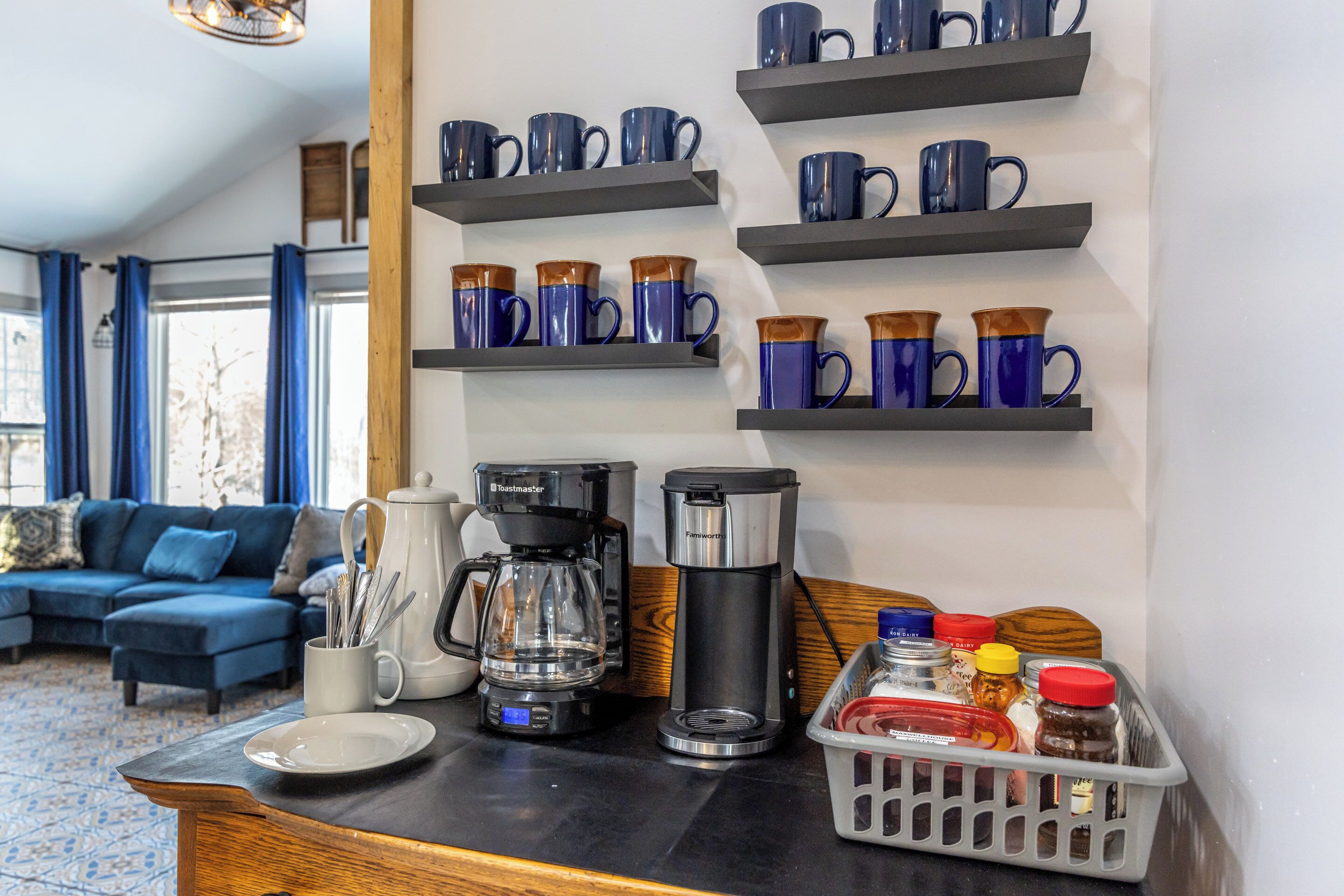 Una sala de estar moderna con un sofá, una mesa de centro y estanterías con tazas azules. Hay dos cafeteras y una cesta de condimentos.