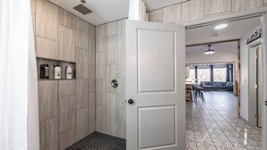 Een moderne badkamer met een douche, betegelde wanden en een deur naar een woonruimte met een bank en een eettafel.