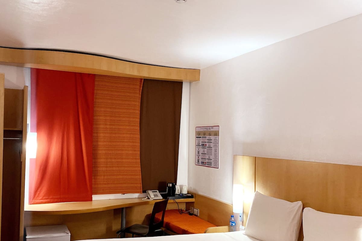 A hotel room with a bed, a desk, a chair, a telephone, and a wall-mounted information board.