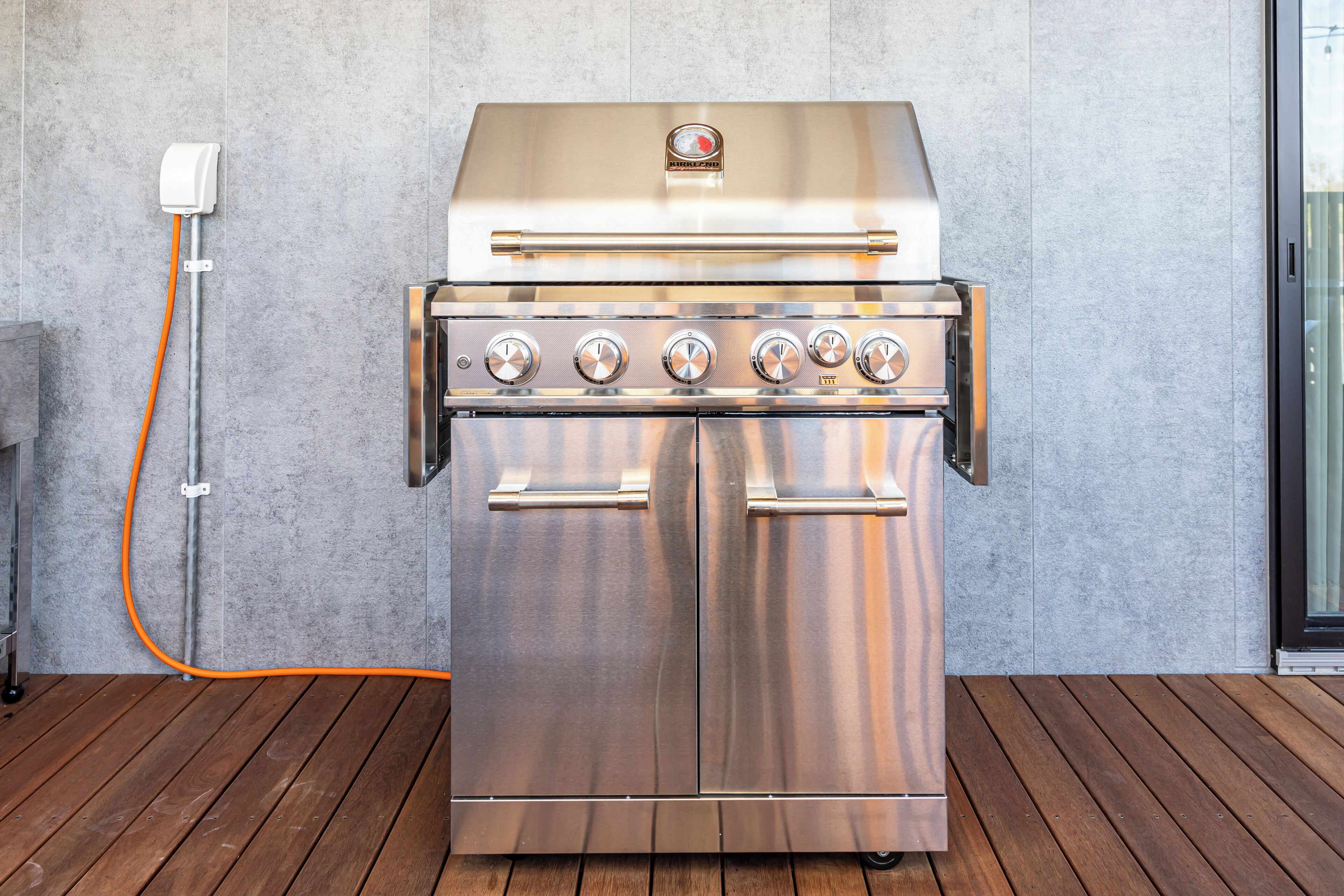 A stainless steel outdoor grill with a control panel and a hose, set against a concrete wall on a wooden deck.