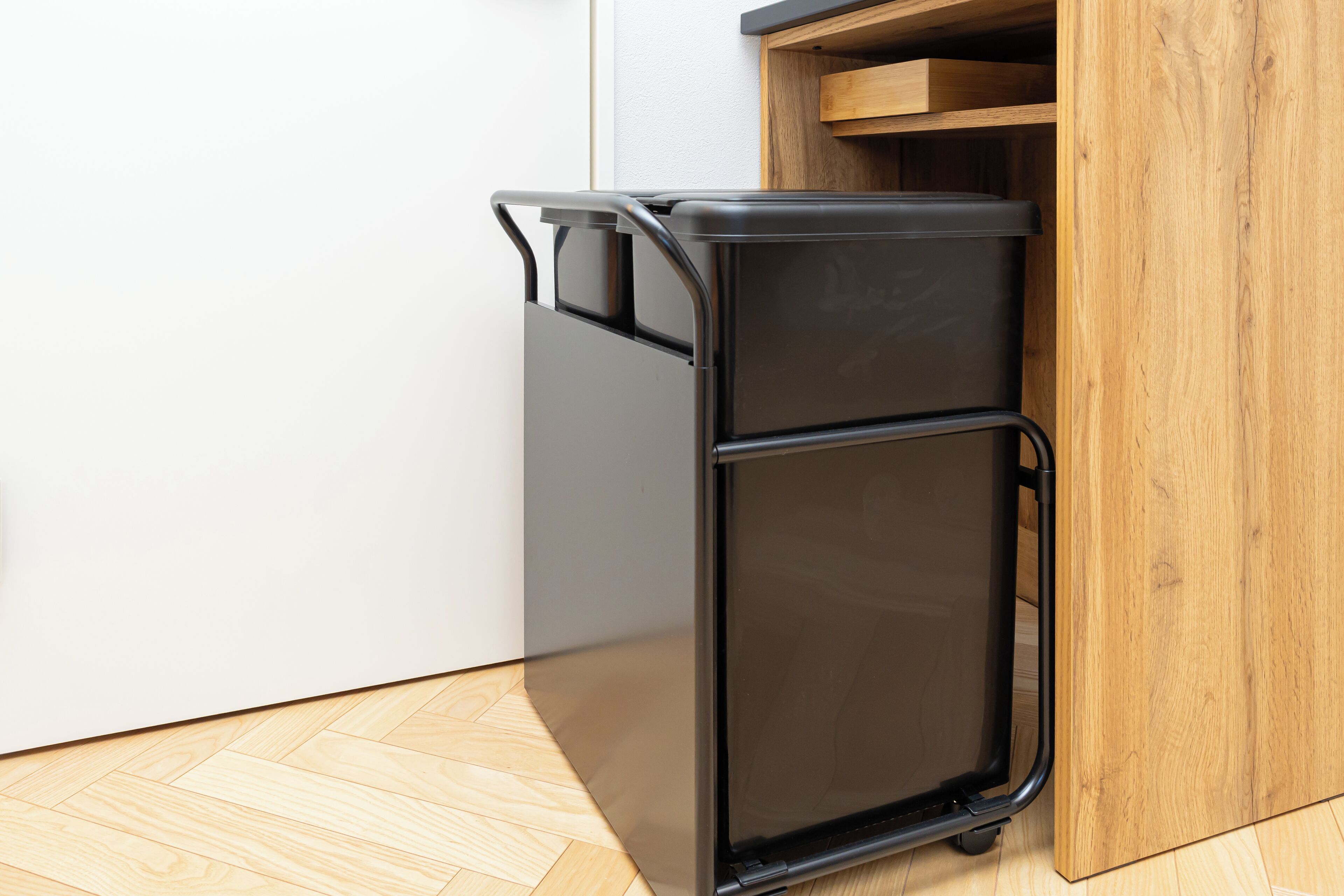 Un meuble de rangement compact, noir, muni de roulettes et doté d’une porte coulissante, placé dans un coin, à côté d’une armoire en bois et d’un mur blanc.