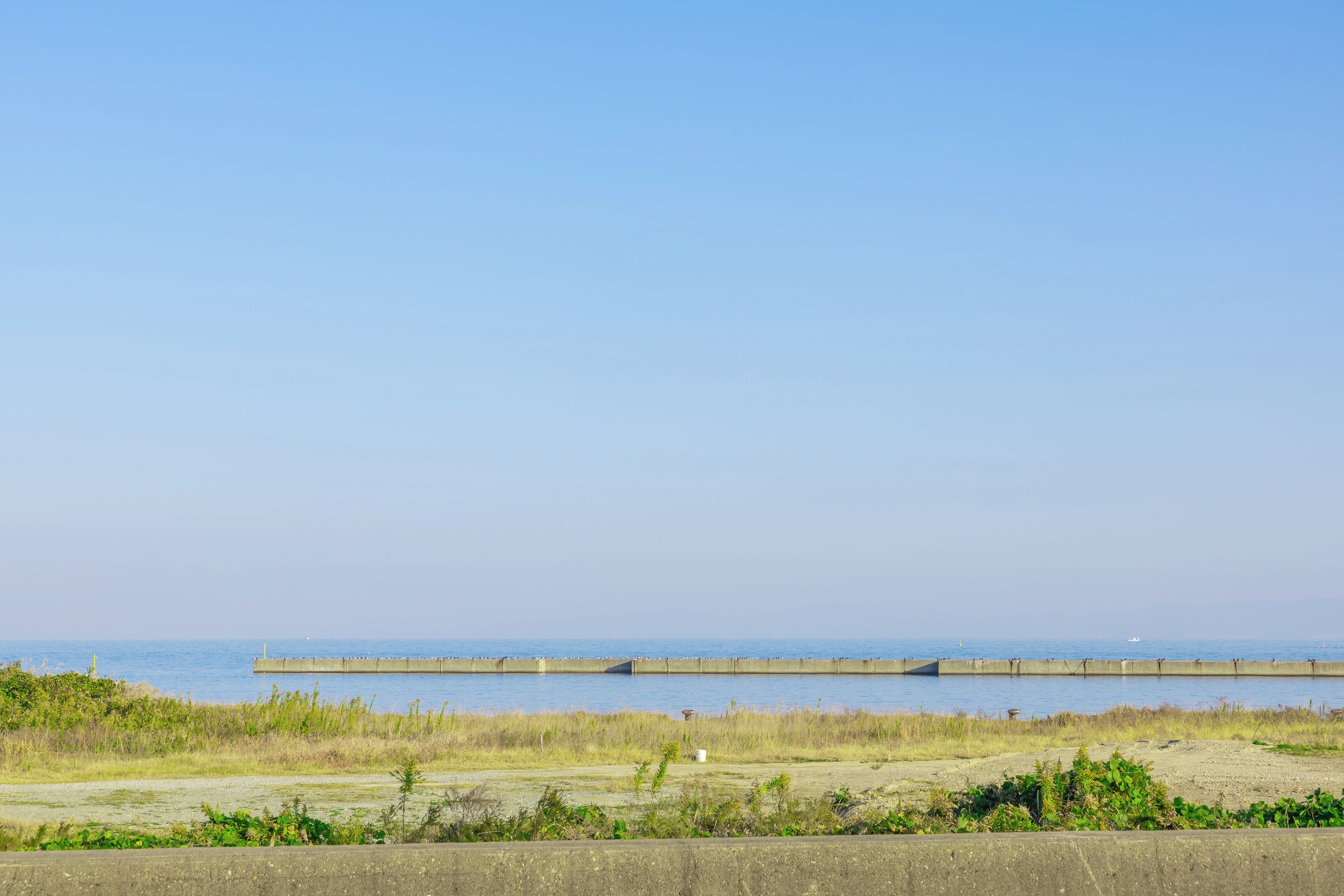 A coastal landscape with a concrete breakwater extending into the sea, a sandy beach in the foreground, and a clear sky.