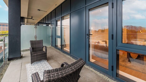 Een modern balkon met rieten stoelen, een patroonkleed, grote glazen deuren en uitzicht op de stad.