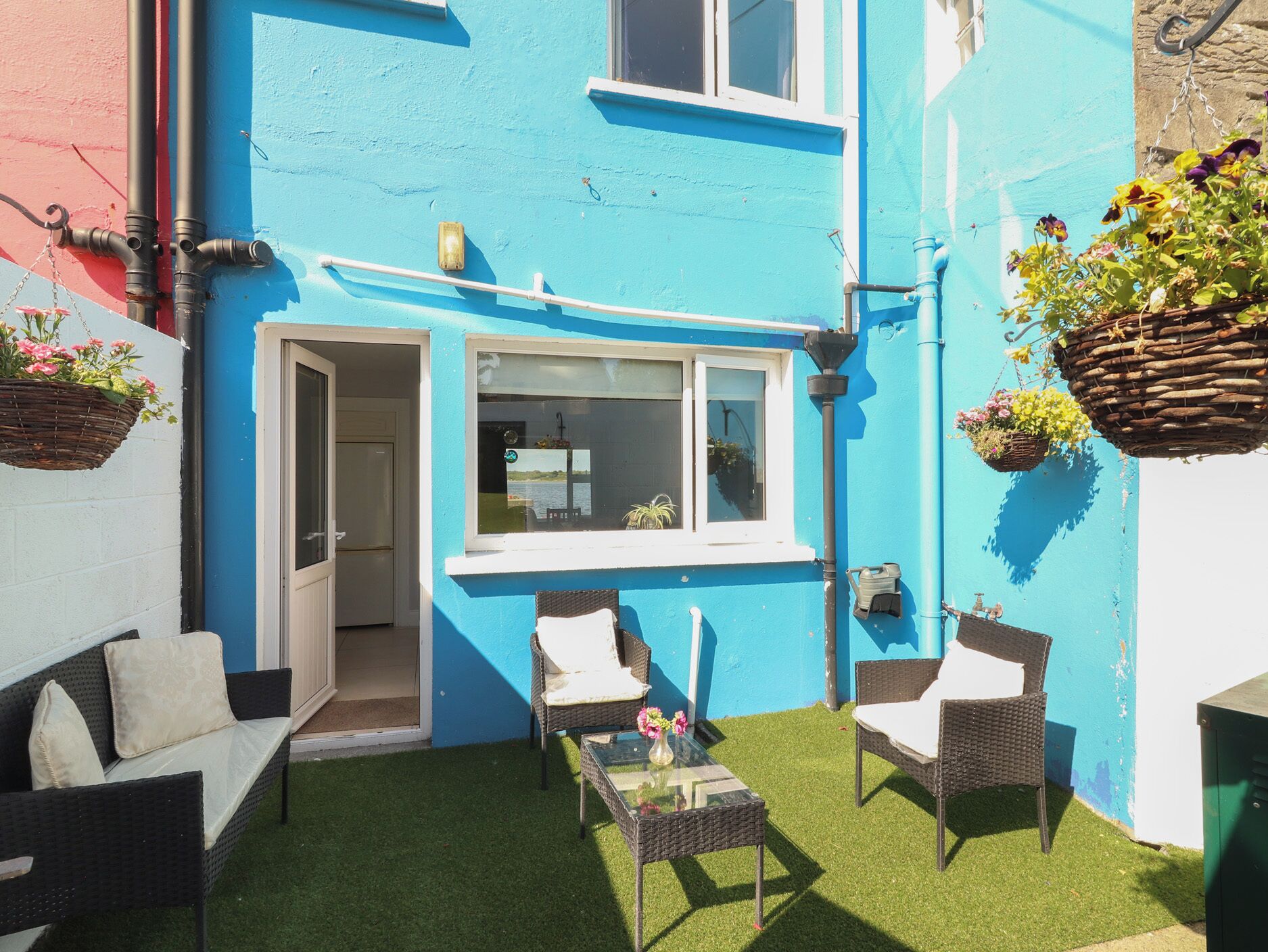 A blue house with a small patio area, two wicker chairs, a table, and hanging flower baskets.