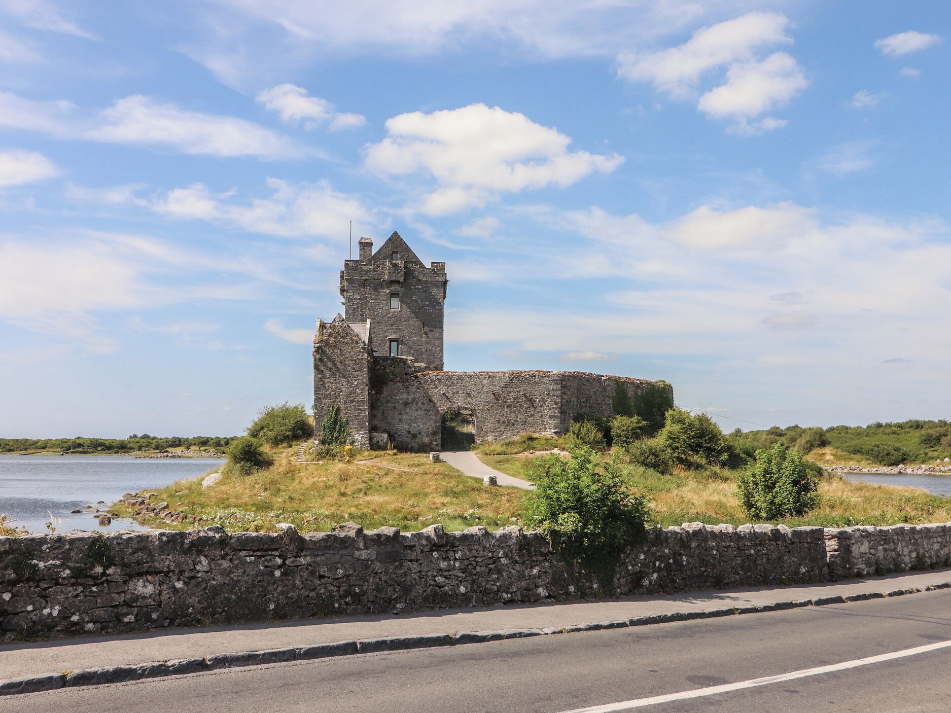 A historical stone structure with a tower, situated by a body of water, accessible by a paved path.