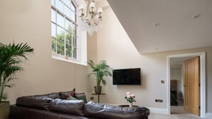 Un salon moderne avec un canapé en cuir marron, une grande fenêtre, un lustre, une plante en pot et un téléviseur à écran plat fixé au mur.