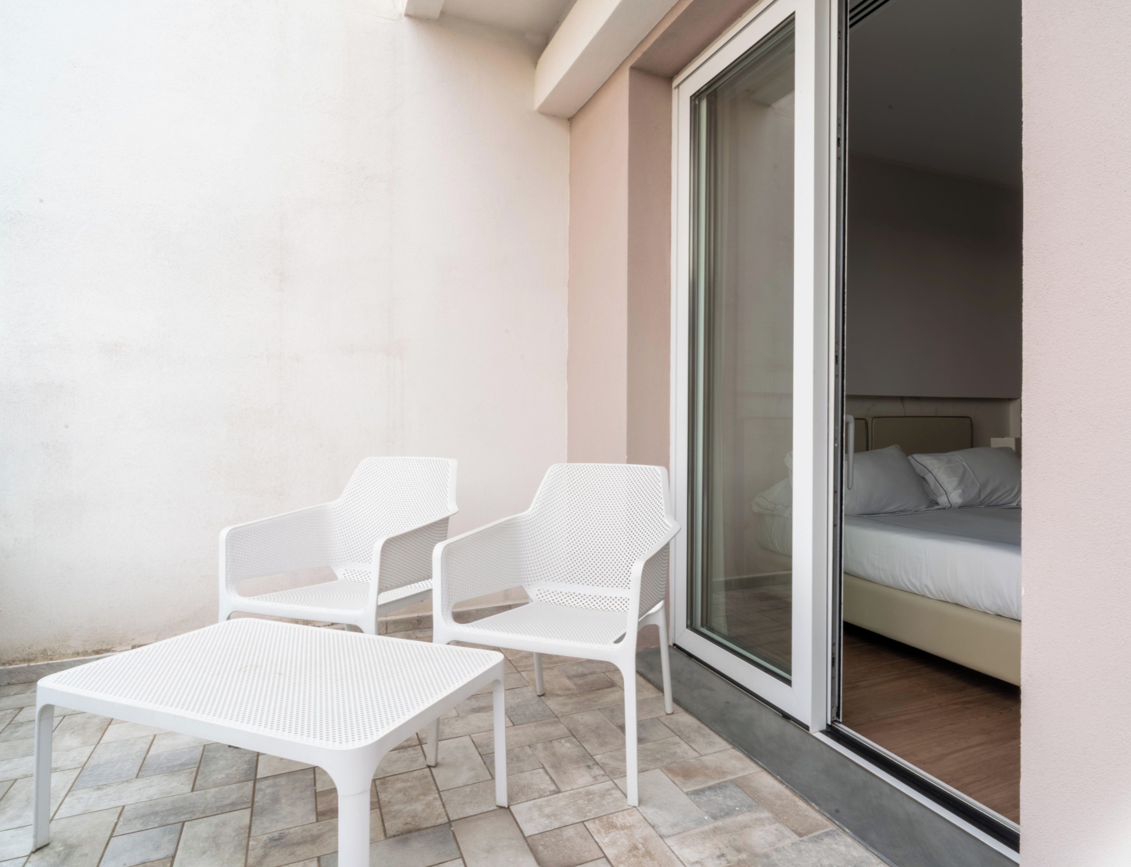 Ein Balkon mit zwei weißen Stühlen und einem Tisch, eine Glastür zum Schlafzimmer mit einem Bett und Kissen.