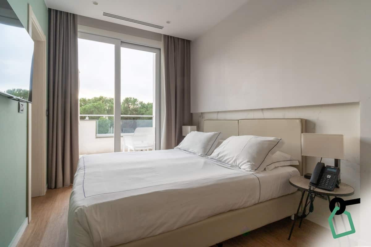 A hotel room with a large bed, a bedside table with a lamp, and a view of the outdoors through a sliding door.