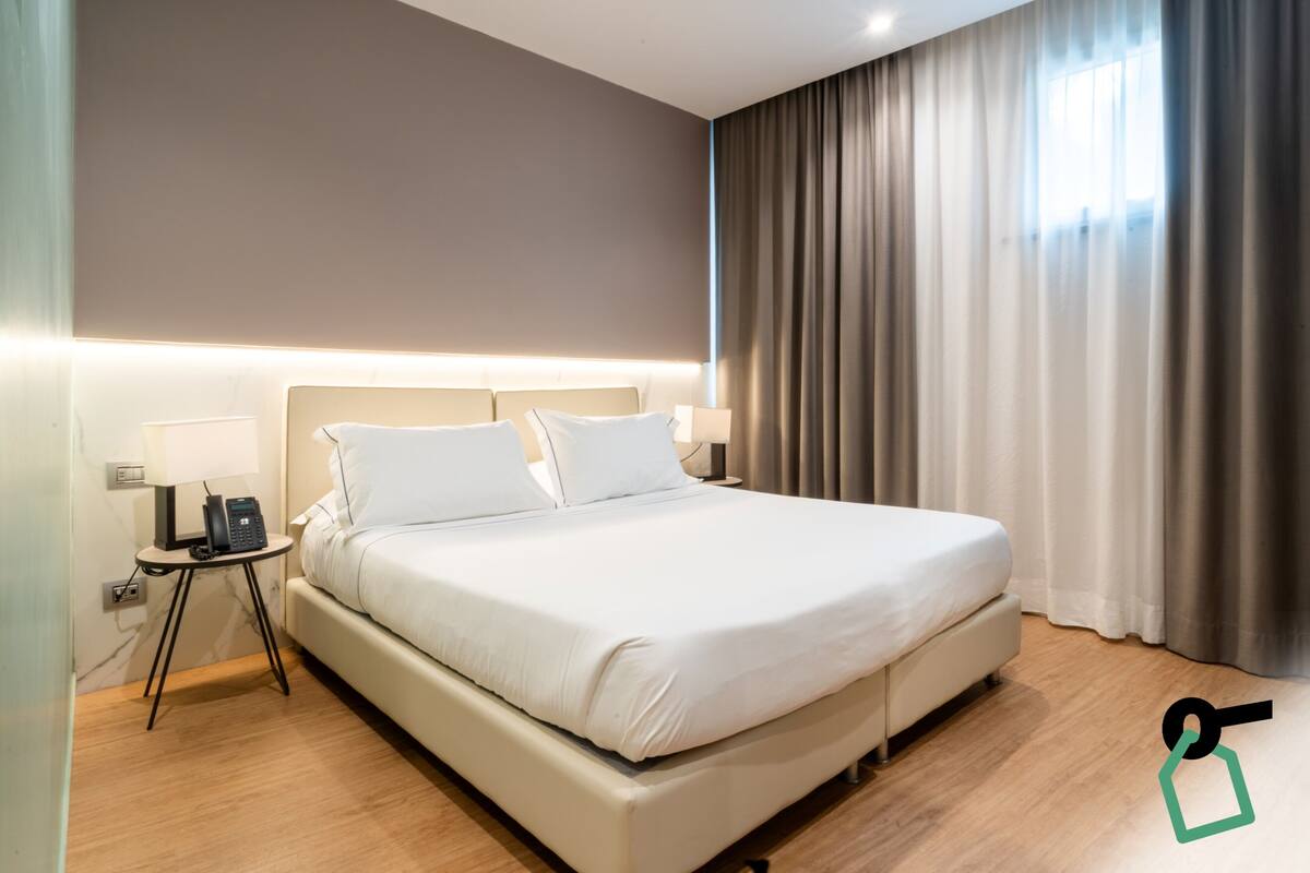 A modern hotel room with a large bed, bedside lamps, a telephone, and a view of the outside through curtains.