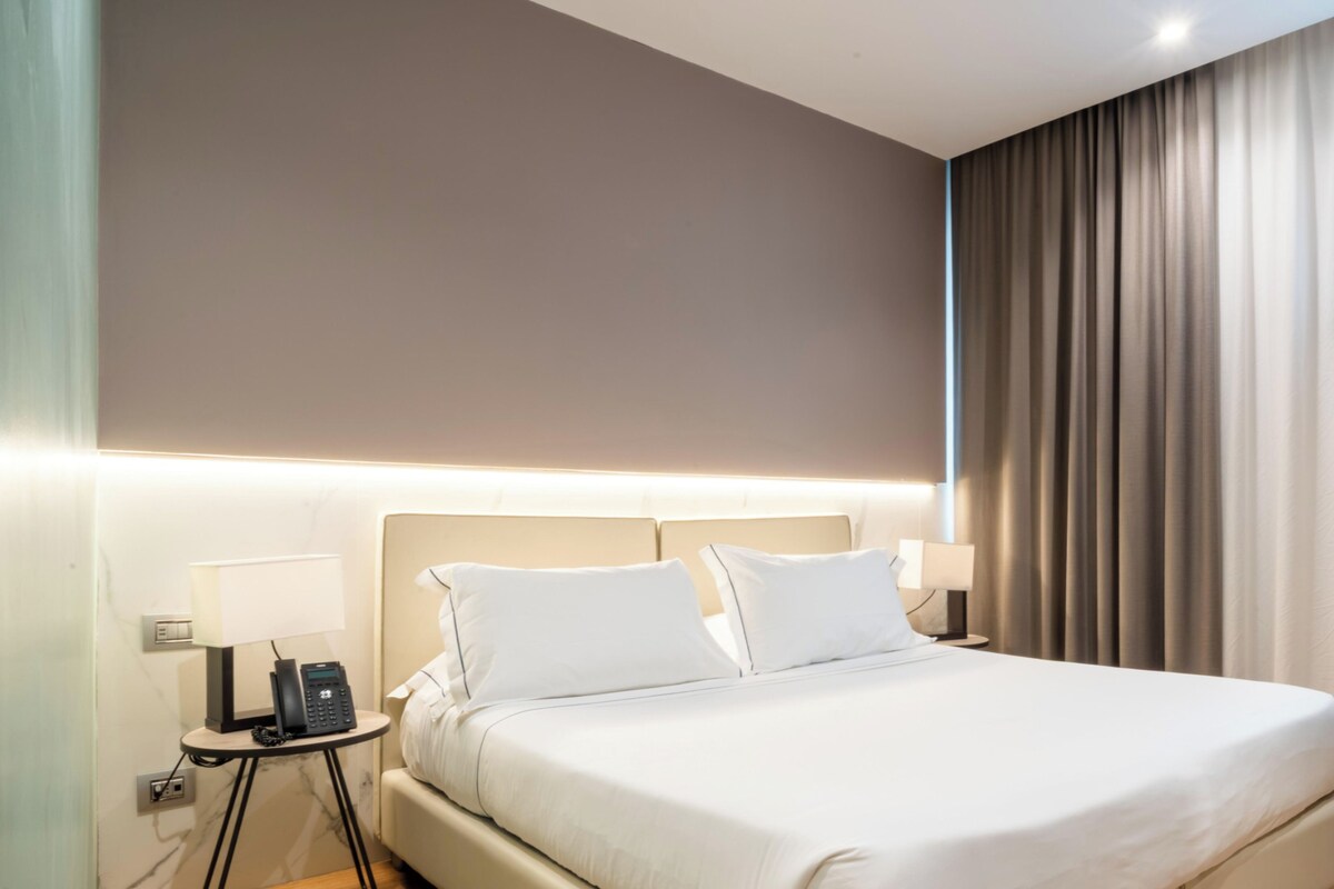 Habitación de hotel moderna con una cama grande, lámparas de noche, un teléfono y vistas al exterior a través de cortinas.