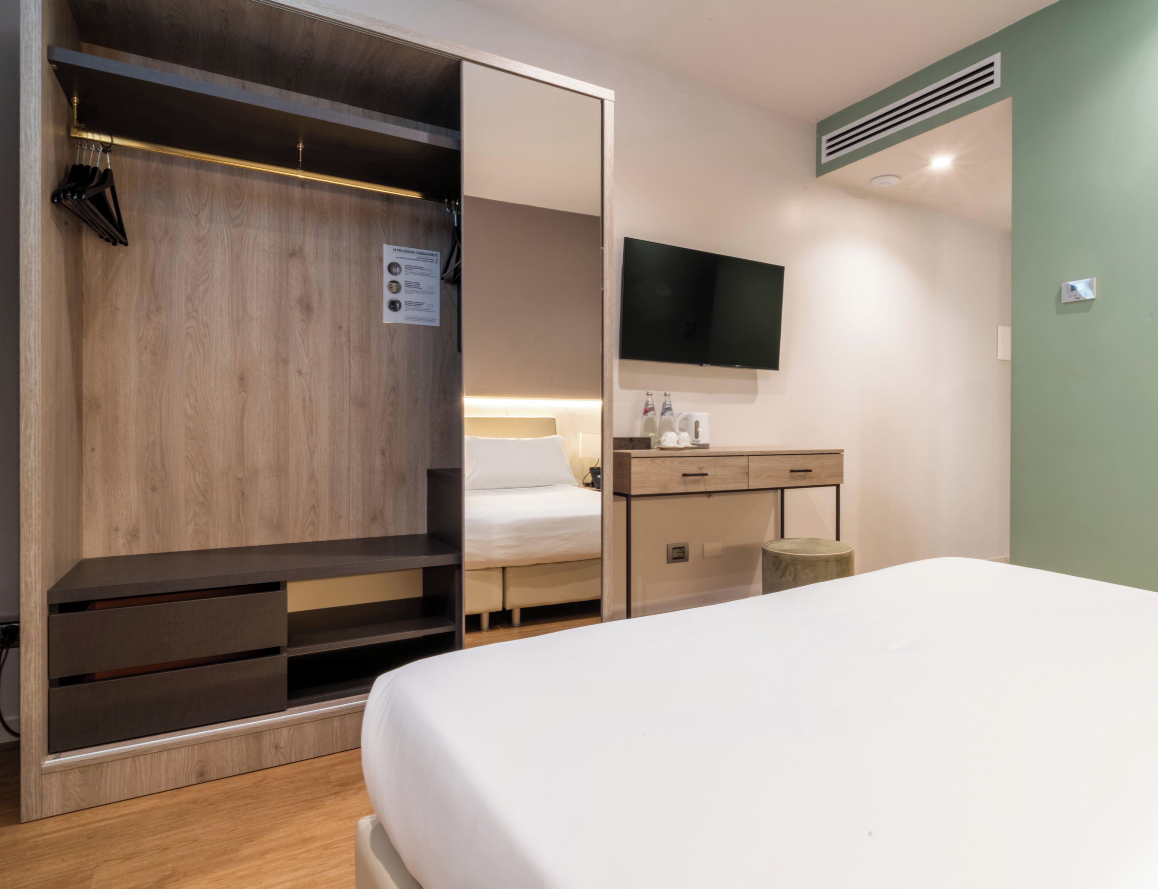 A hotel room with a bed, a wardrobe with a mirror, a desk with a flat-screen TV, and a small stool.