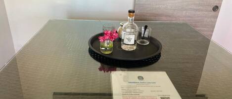 Una mesa de vidrio con una bandeja que contiene una botella etiquetada como "LABAT 50", un vaso con flores rosadas y un pequeño recipiente con líquido verde. Un documento con información del hotel está colocado sobre la mesa.