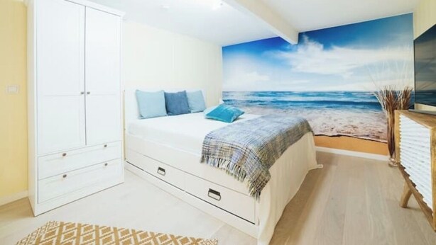 Een slaapkamer met een bed, een kledingkast, een strandschildering en een kleine tafel met een plant.