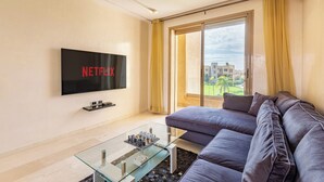 A modern living room with a large flat-screen TV mounted on the wall, a glass coffee table, a dark-colored sofa, and a view of a cityscape through a large window. - Cozy apartment with pool in Argan, 3 bedrooms (Marrakesh)