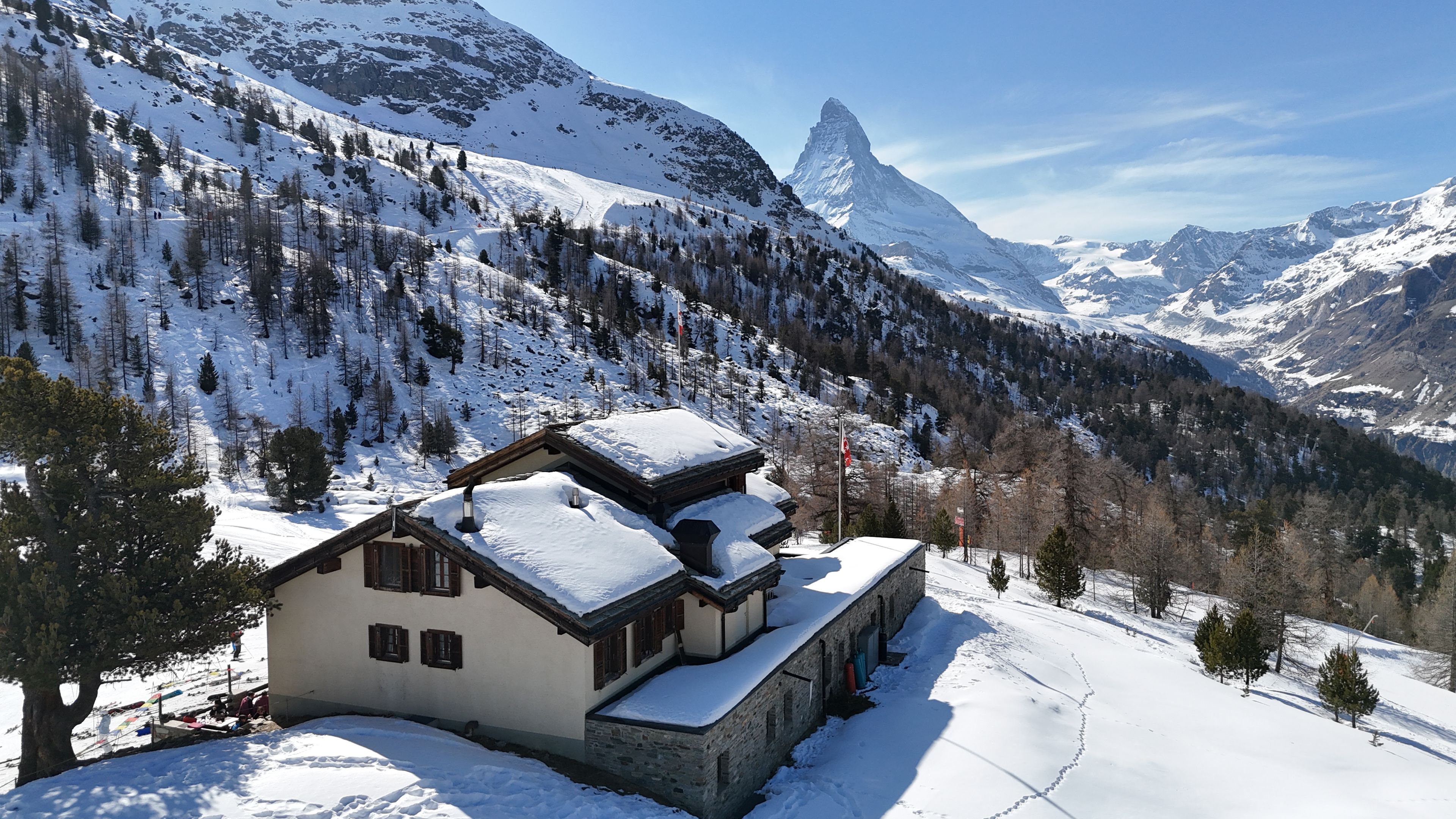 Een chalet met een met sneeuw bedekte dak, gelegen in een bergachtig gebied met een markante bergtop op de achtergrond.