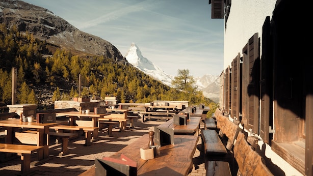Outdoor wooden tables and benches arranged in rows, with a mountainous backdrop and a building with wooden shutters. - Grünsee Mountain Lodge (Zermatt)