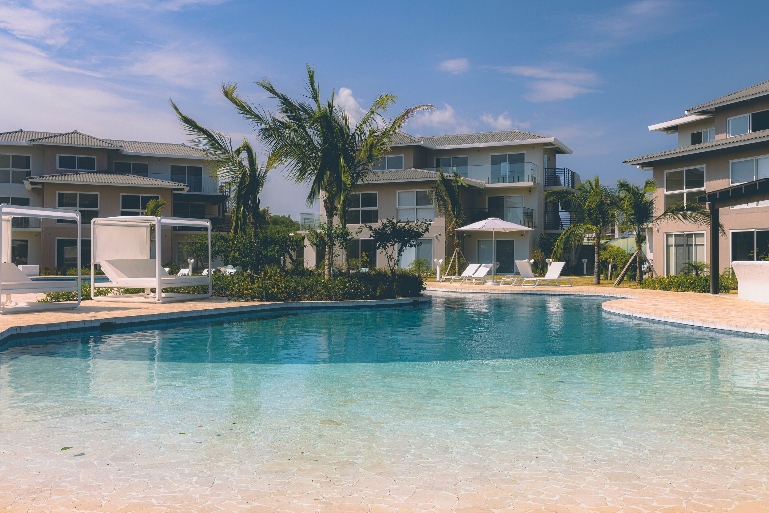 Una piscina con sillones y sombrillas, rodeada de palmeras y edificios modernos.
