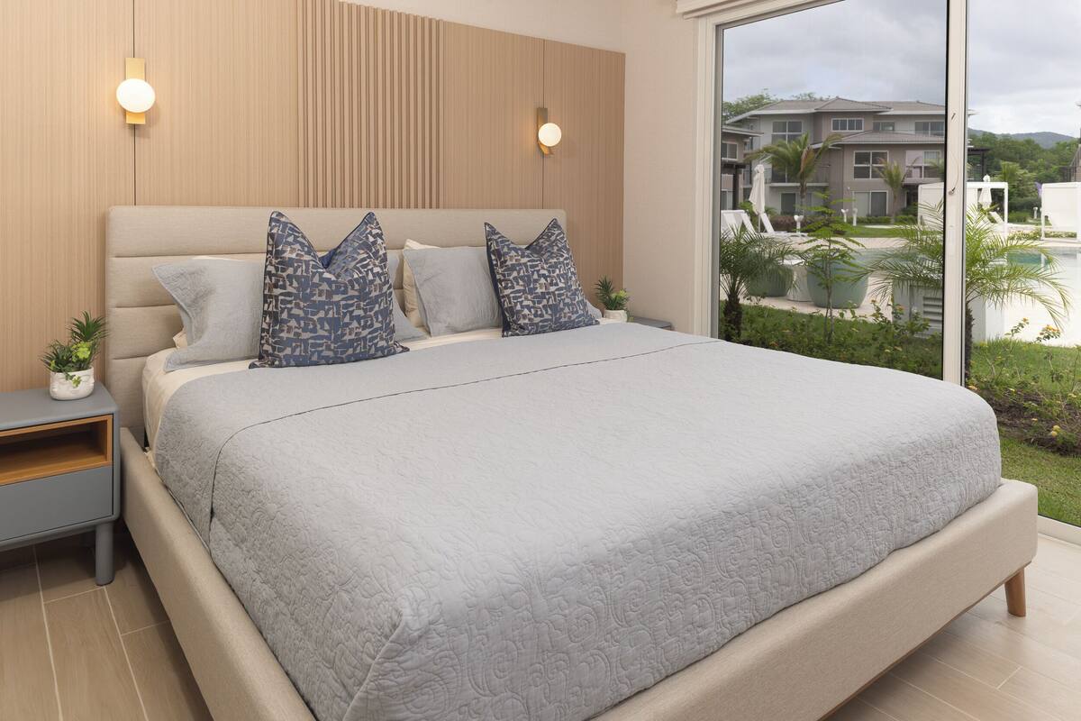 Un dormitorio moderno con una cama grande, dos mesitas de noche, una planta pequeña y vistas a una zona residencial a través de un ventanal amplio.