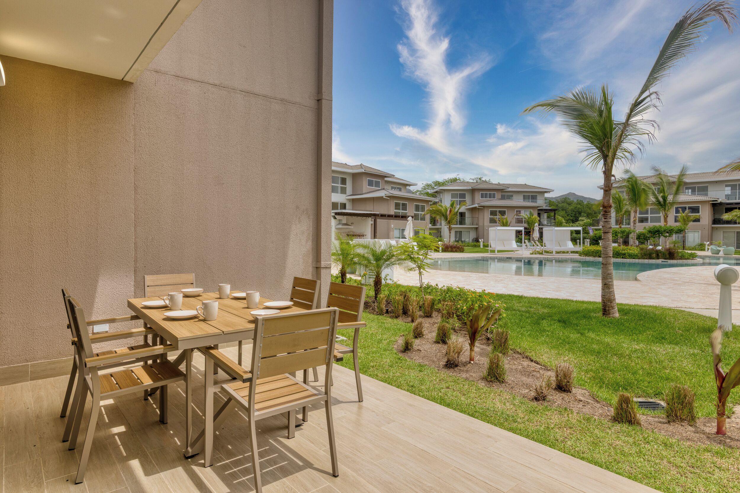 A patio with a wooden table and chairs, a pool, and a view of residential buildings.