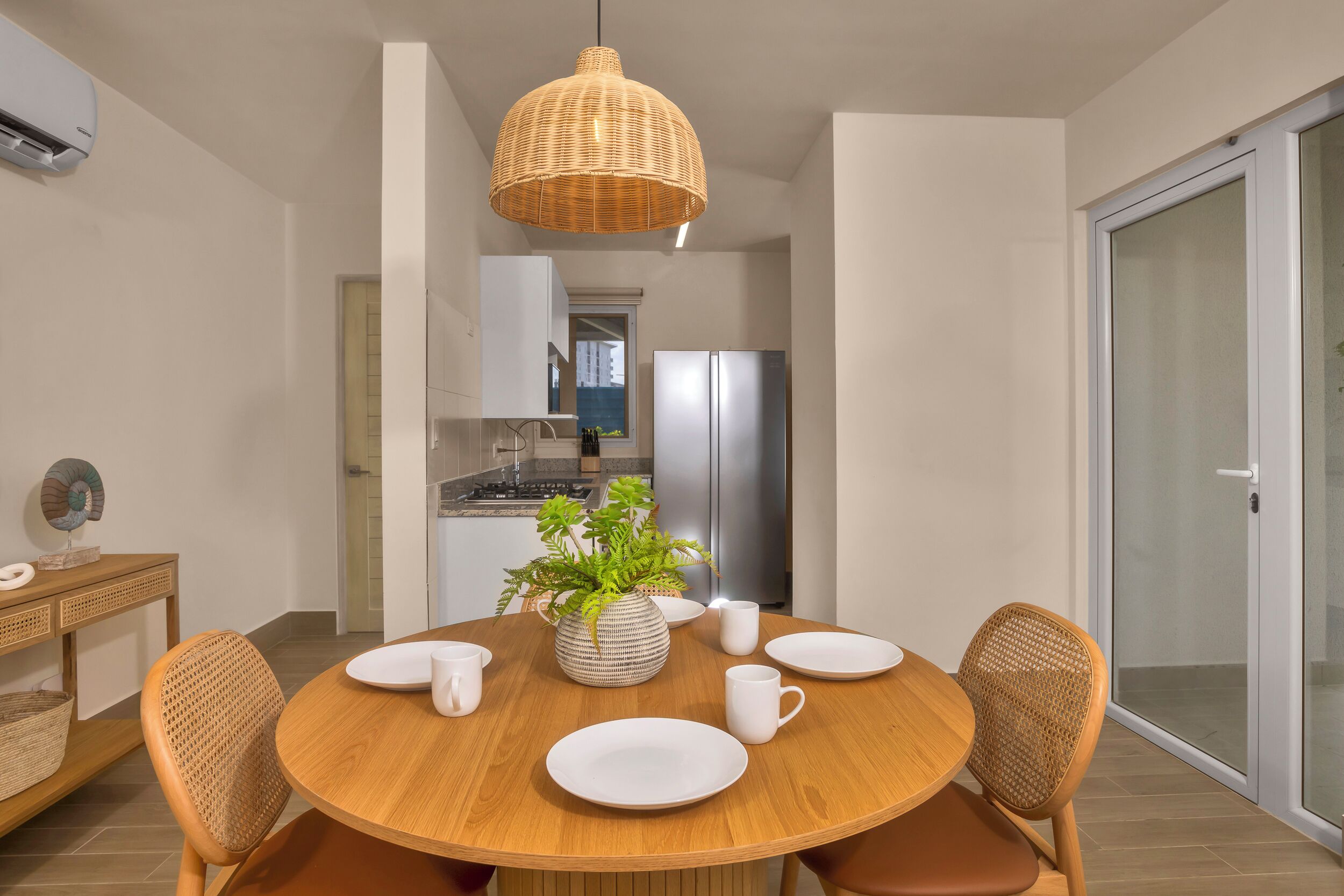A modern dining area with a round wooden table, white plates, and matching chairs. A woven pendant light hangs above, and a potted plant adds greenery.