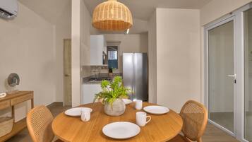 A modern dining area with a round wooden table, white plates, and matching chairs. A woven pendant light hangs above, and a potted plant adds greenery.
