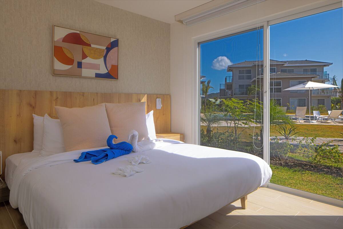 Una cama grande con ropa de cama blanca, un cabecero de madera y vistas a un resort a través de un ventanal.