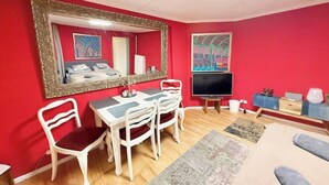 Quarto com parede vermelha, um espelho grande, uma cama, uma televisão, uma mesa de jantar com cadeiras e um sofá.