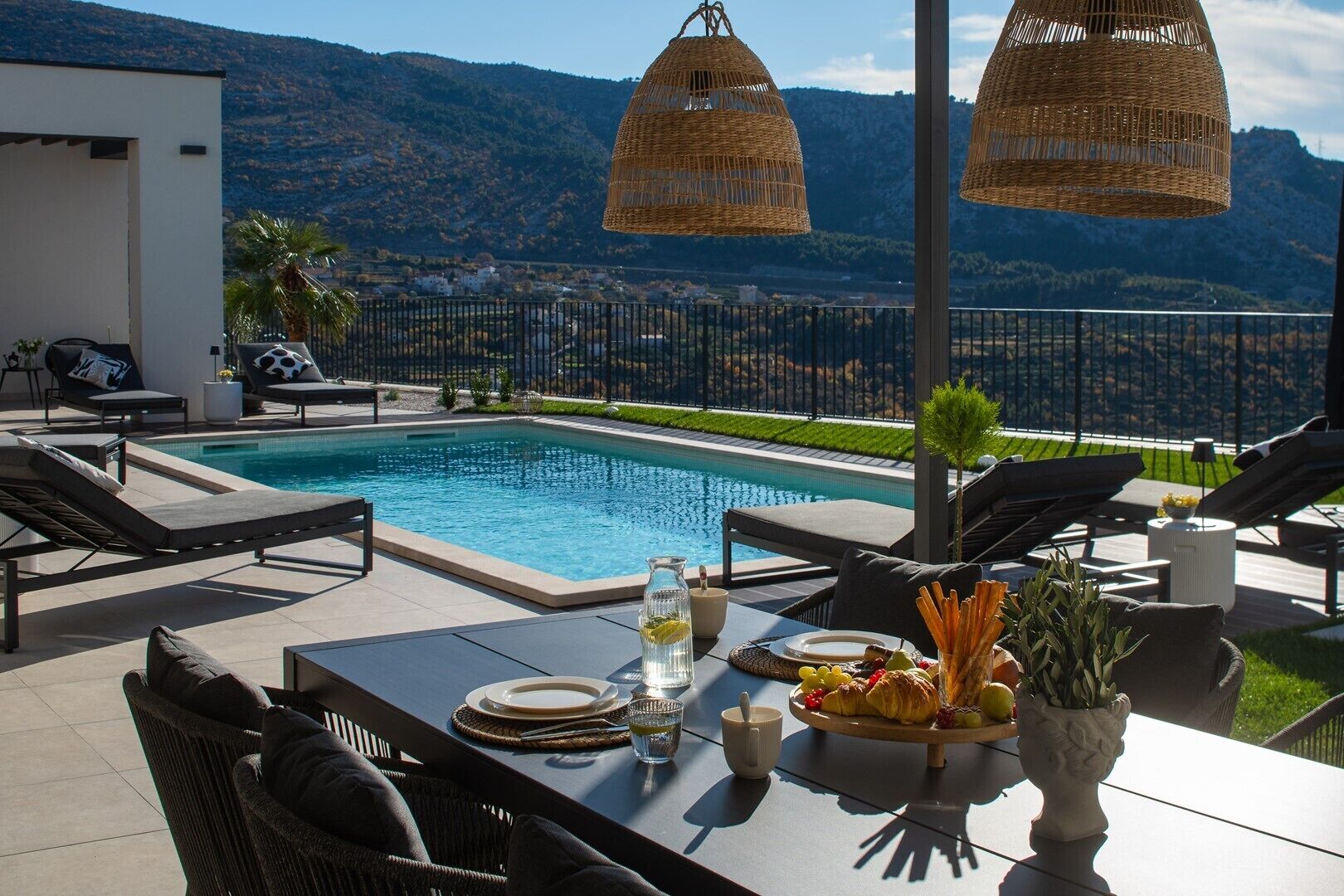 A poolside area with a table set for dining, lounge chairs, and a view of mountains.