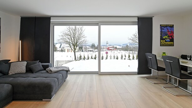 Ein modernes Wohnzimmer mit einem großen Schiebe-Glaskamin, der auf eine verschneite Außenanlage führt, ein graues Sofa und ein Essbereich mit Stühlen und einem Tisch.