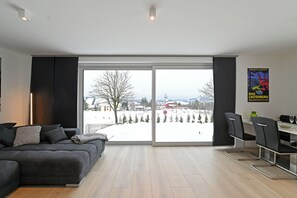 A modern living room with a large sliding glass door leading to a snowy outdoor area, a grey sofa, and a dining area with chairs and a table. - HarZeit 1E Vacation Home with Sauna and Adventure Bed (Sankt Andreasberg)