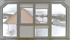 Ein Zimmer mit großem Fenster mit Blick auf eine verschneite Landschaft, darunter ein Holzhaus und ein eingezäunter Bereich.