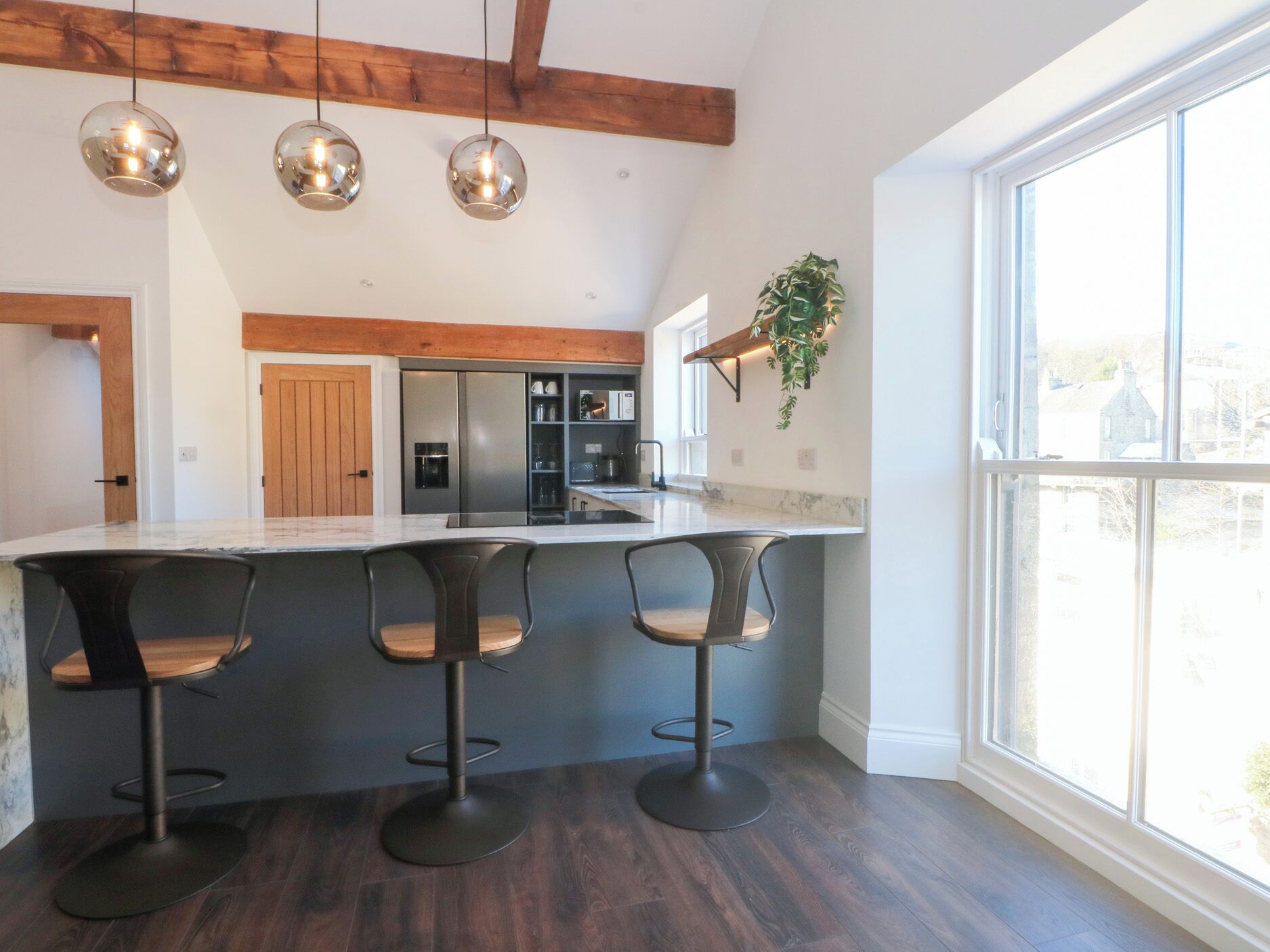 A modern kitchen with a marble countertop, three bar stools, a built-in refrigerator, and pendant lighting.