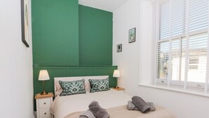 A bedroom with a green headboard, two bedside lamps, a bed with a beige blanket and two rolled towels, a small white nightstand, and a window with blinds. - The Penthouse at 86 (Rossendale)