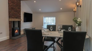A dining area with a brick fireplace, a TV mounted on the wall, and a window with blinds. - The Penthouse at 86 (Rossendale)