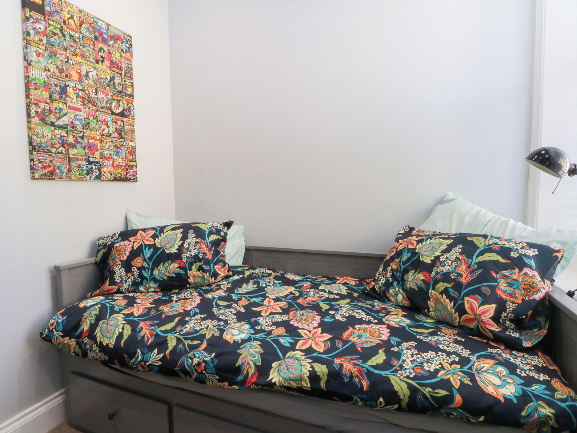 A bedroom with a bed, floral-patterned bedding, a wall art piece, and a bedside lamp.