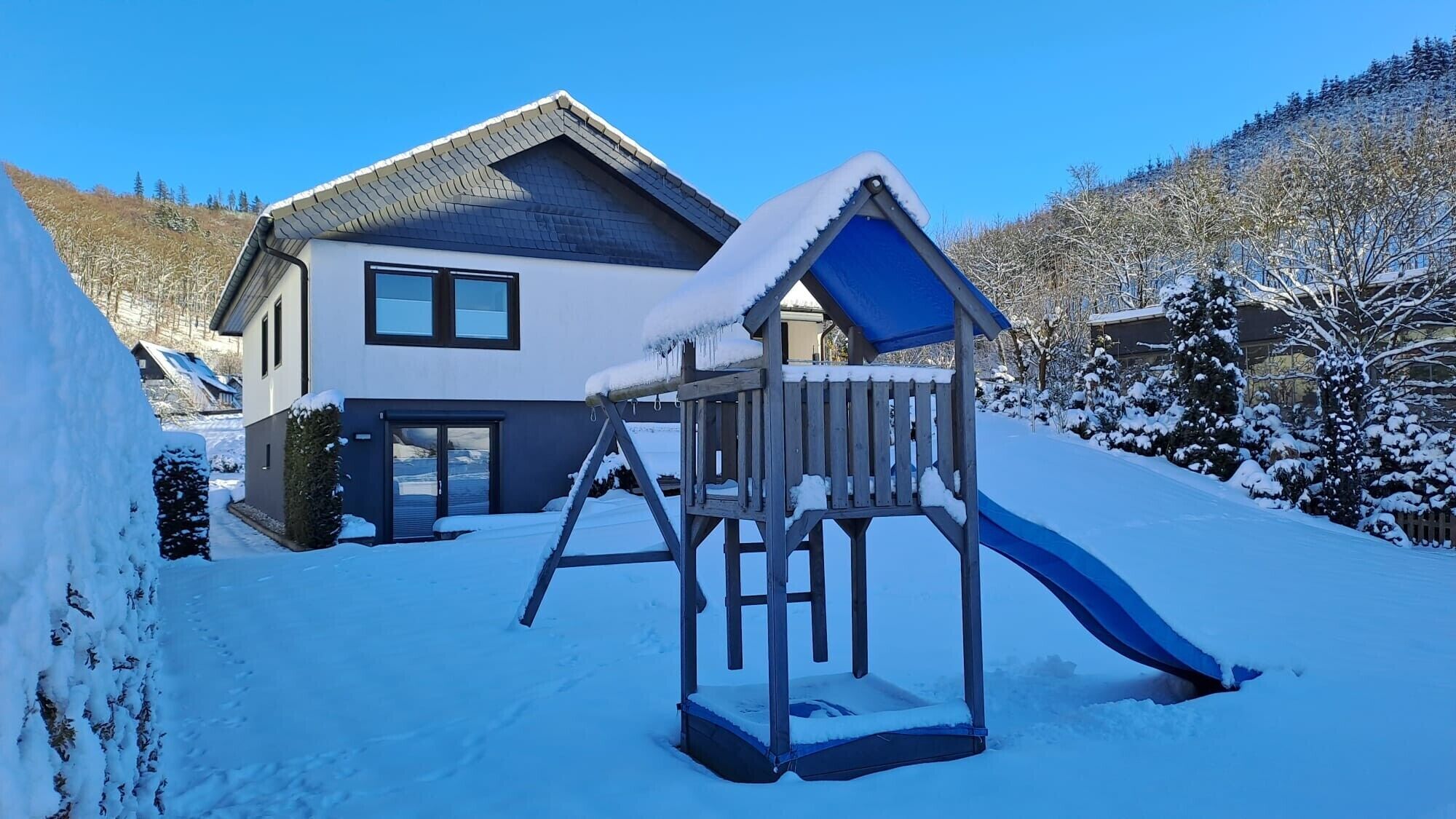 A snow-covered playground with a slide, a house with a sloped roof, and a snowy hillside in the background.