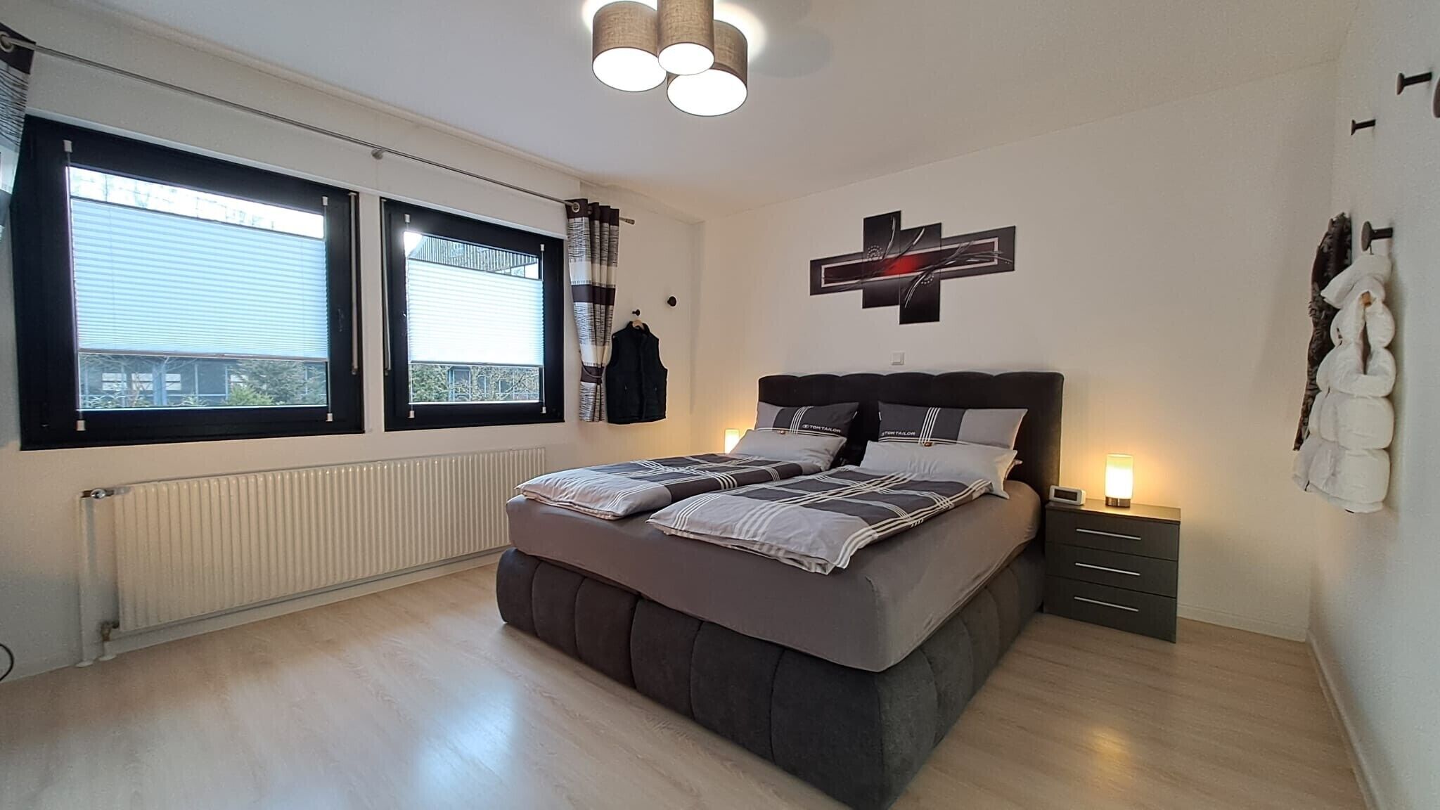 A bedroom with a bed, a radiator, a window with blinds, a wall-mounted cross, and a bedside table with a lamp.