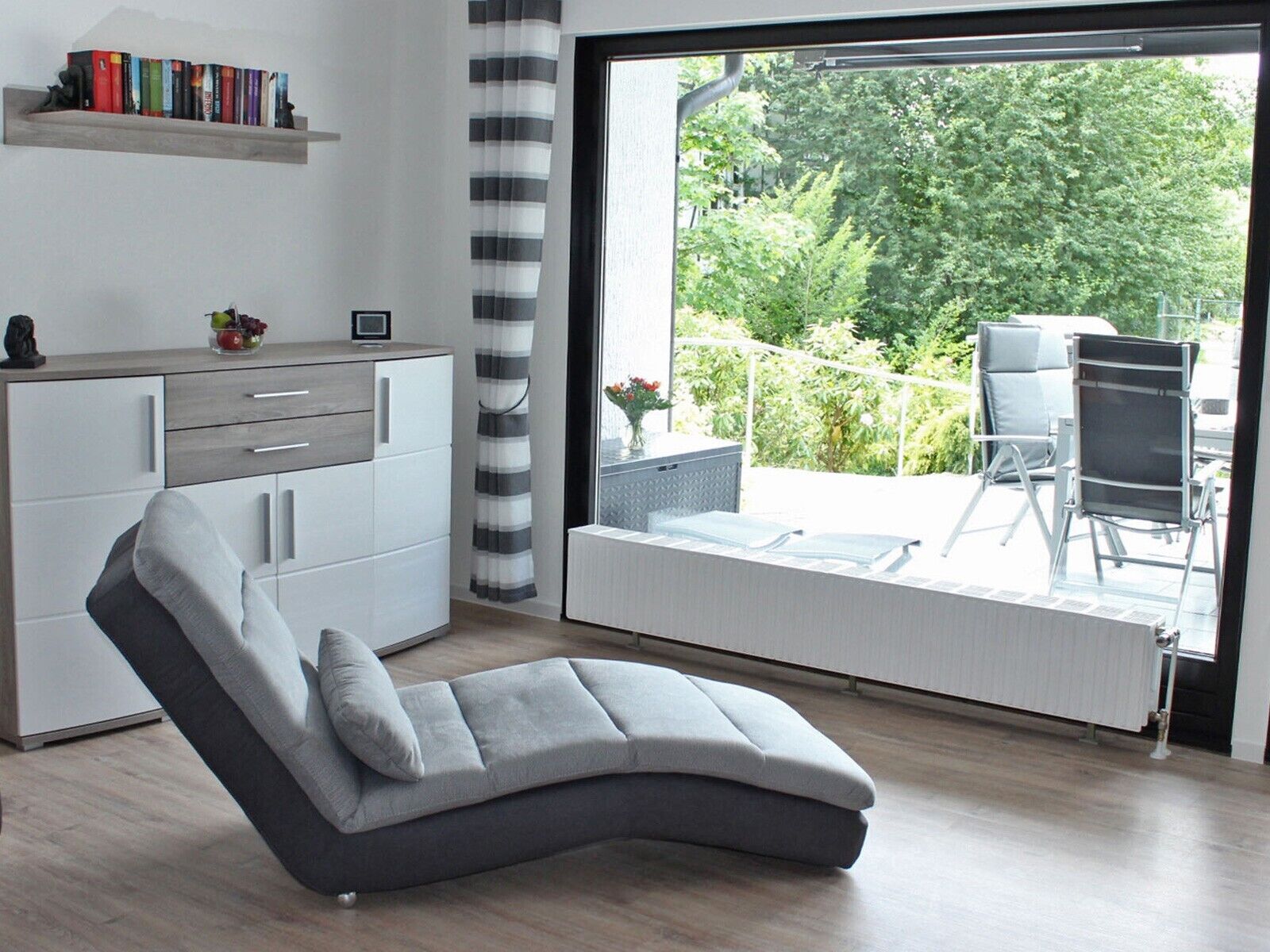 A modern living room with a grey sofa, a white cabinet, a bookshelf, and a large sliding glass door leading to an outdoor patio with a chair and table.