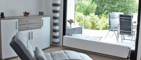 A modern living room with a grey sofa, a white cabinet, a bookshelf, and a large sliding glass door leading to an outdoor patio with a chair and table.
