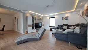 A modern living room with a grey sofa, a reclining chair, a TV, and a bookshelf. - Holiday Home Am Rarbach, 140 m² – By the Village Park and Stream, Large Terrace with Mountain View (Schmallenberg)