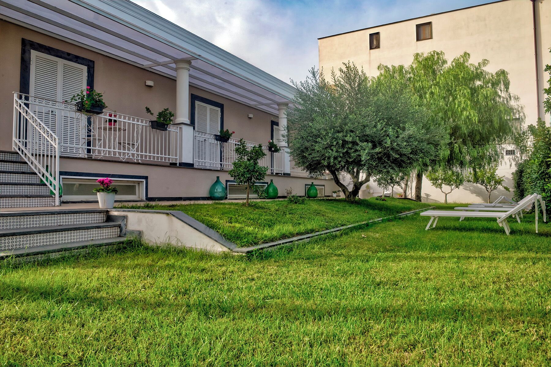 A building with a covered outdoor area, steps leading to a balcony, and a grassy lawn with a bench.