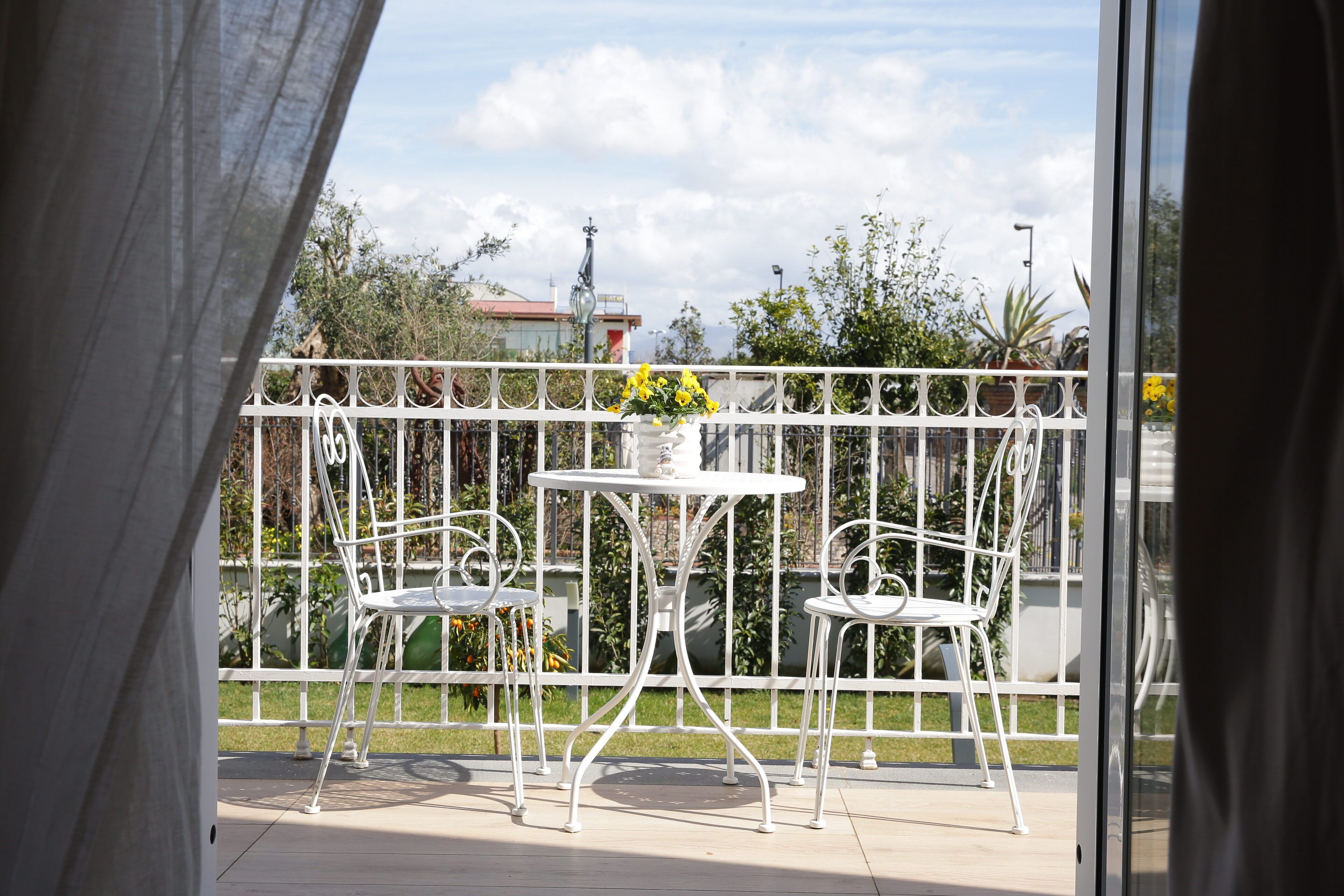 A balcony with white metal chairs and a table, a white metal fence, and a view of buildings and trees in the background.