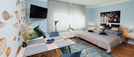 Une chambre moderne comprenant un lit, des tables de chevet, un bureau avec des chaises, une grande fenêtre avec des rideaux et un téléviseur fixé au mur.