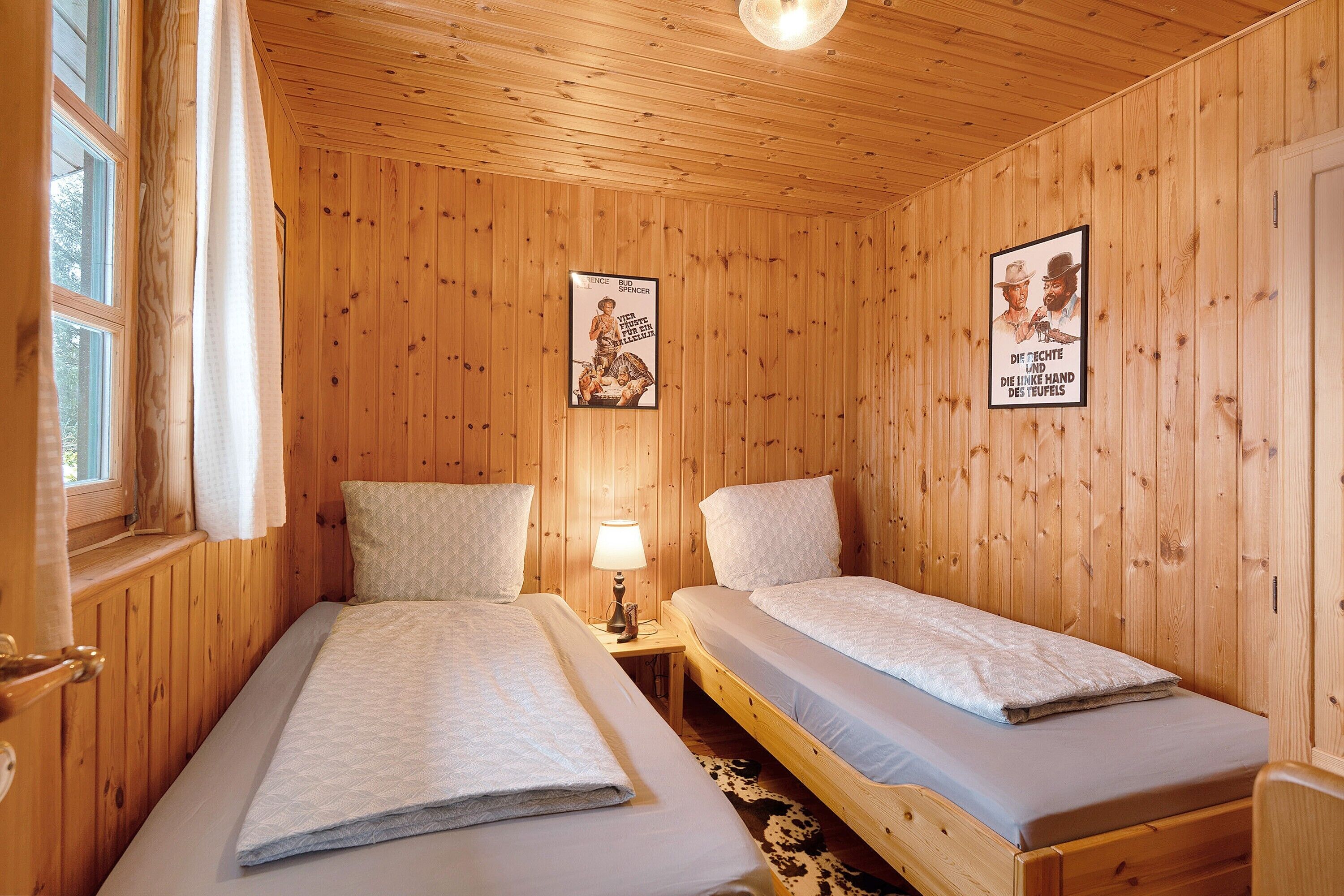 A wooden-paneled room with two beds, a window with curtains, a lamp on a bedside table, and two posters on the wall.
