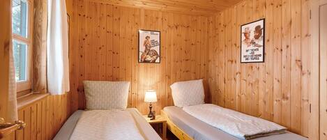 A wooden-paneled room with two beds, a window with curtains, a lamp on a bedside table, and two posters on the wall.