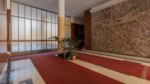 Un hall d’entrée spacieux, doté d’un tapis rouge, de grandes fenêtres à vitres dépolies, d’un mur en pierre et de plantes en pot.