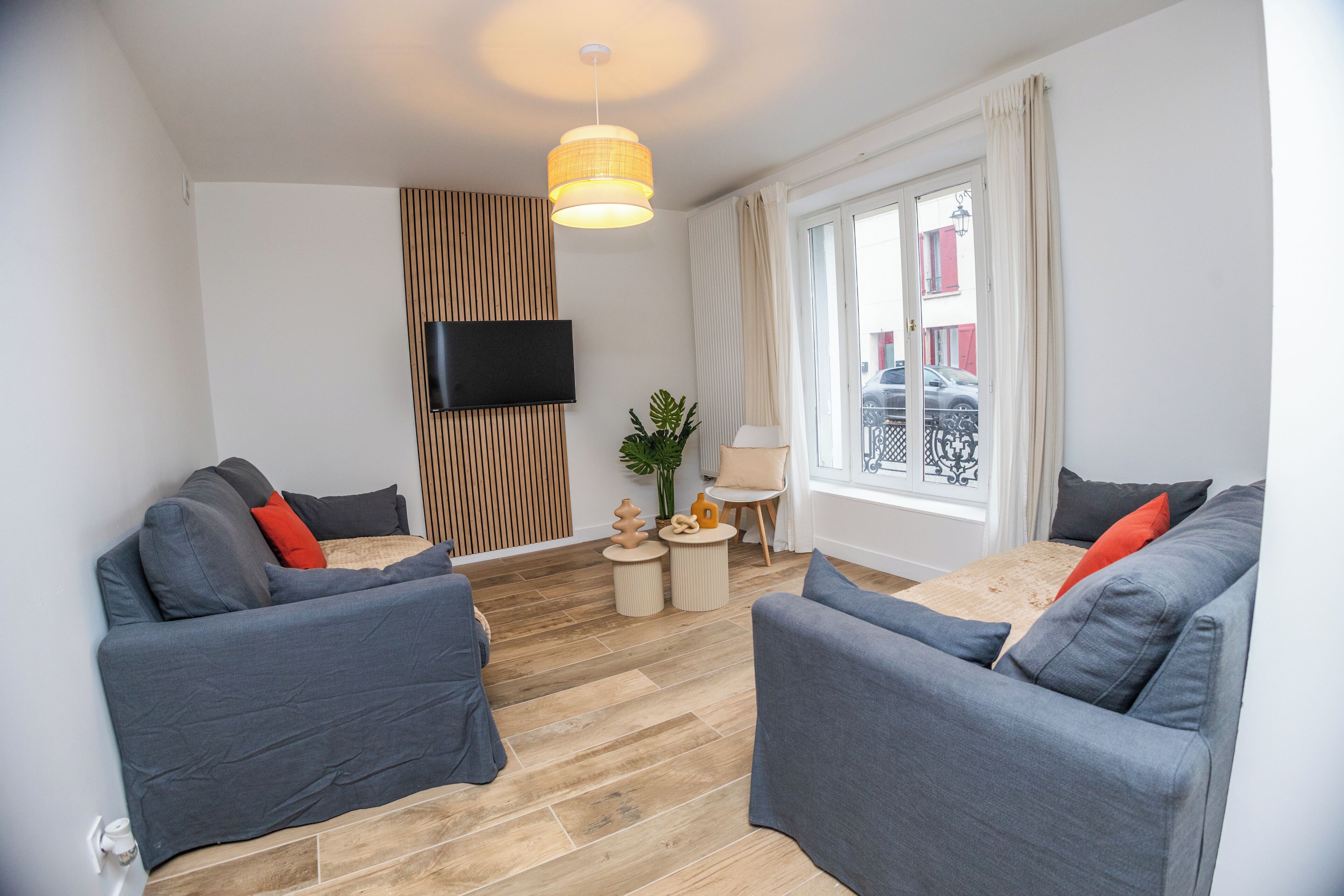 A modern living room with two grey sofas, a wooden floor, a flat-screen TV mounted on the wall, a small round table with a plant, and a window with a view of the street.